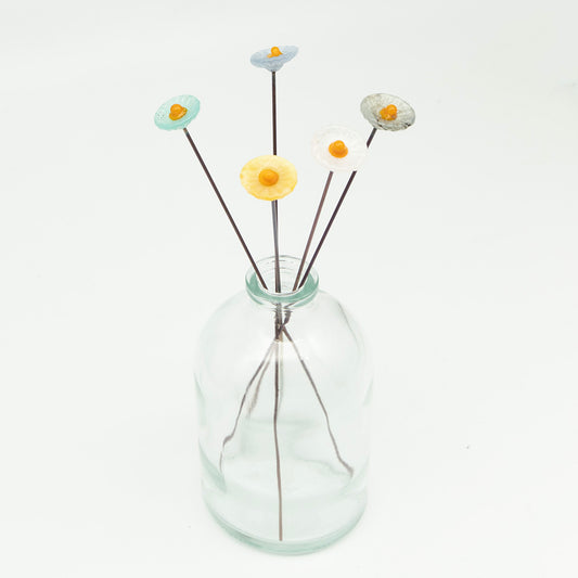 Five pastel glass flowers with metal plant stakes. Flowers are different pastel colours and have yellow centres.  The plant stakes sit in a clear milk bottle vase.