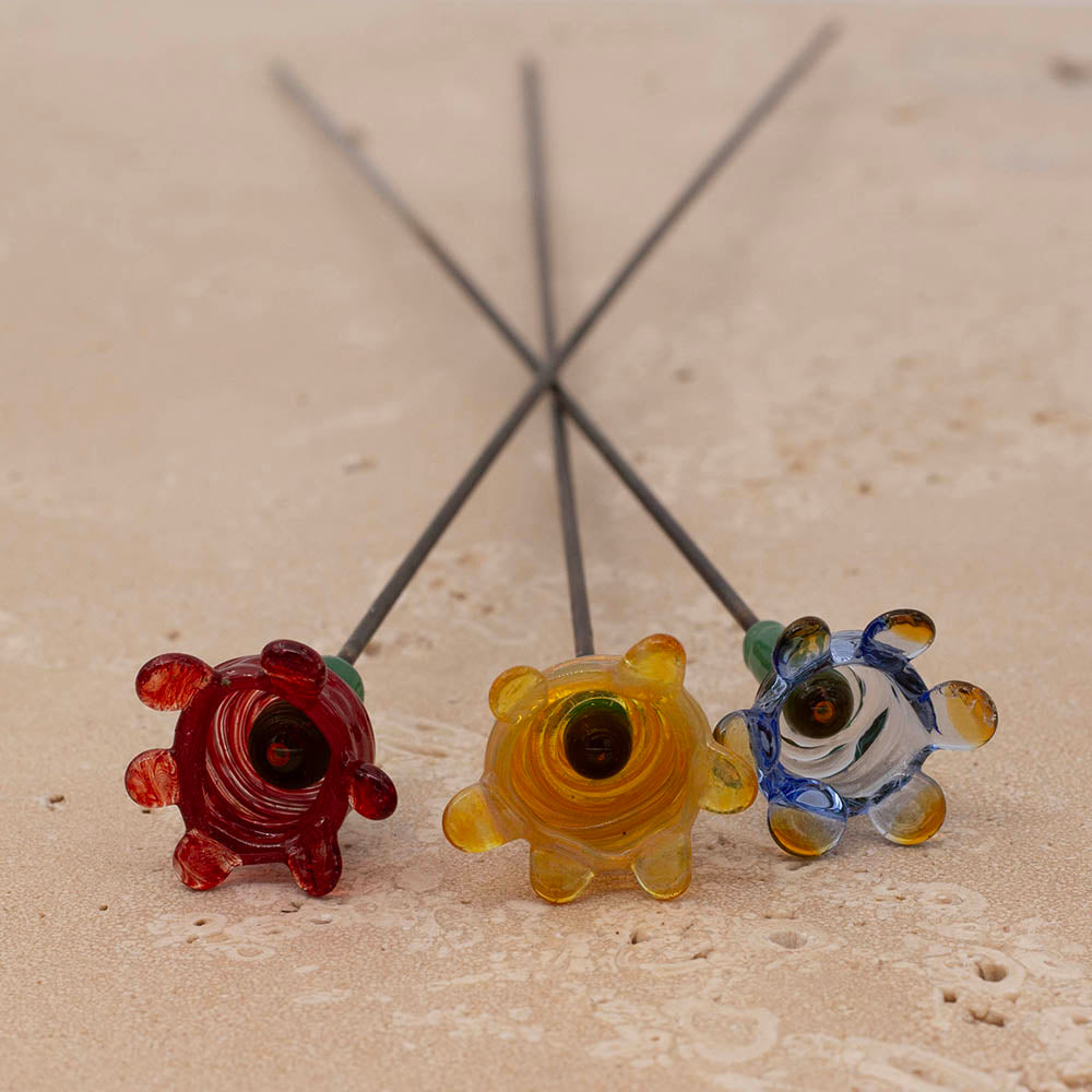 Three bee sippers - flower shapes with curved bottoms to collect water on metal sticks lying on a sandstone tile. The bee sippers are red, yellow and pale blue, and all three have dark centres.