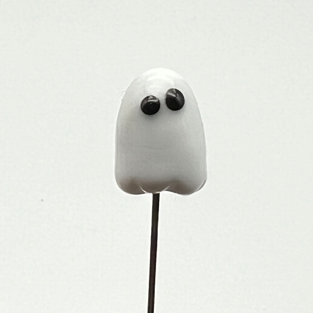 Close up of metal pin with white glass ghost on its head. The ghost has black eyes.