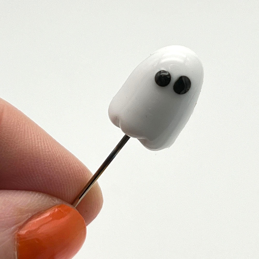 Metal pin topped with a white glass ghost with black eyes. Held between a thumb forefinger.