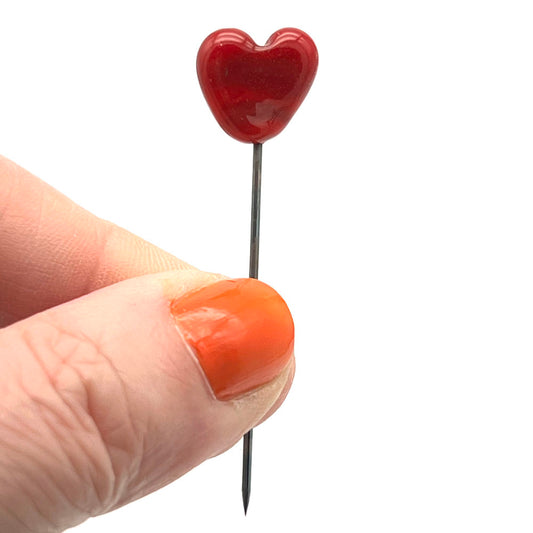 Metal pin with small read heart top shown held between thumb and forefinger. The point of the pin is shown.