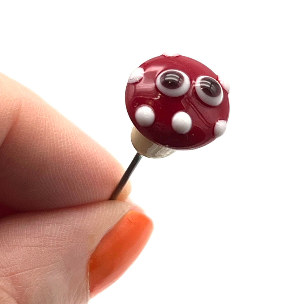 a pin topped with a glass mushroom. The mushroom has a cream stalk and red cap with white dots. It also has two cartoon style eyes. Shown held between a thumb and forefinger.