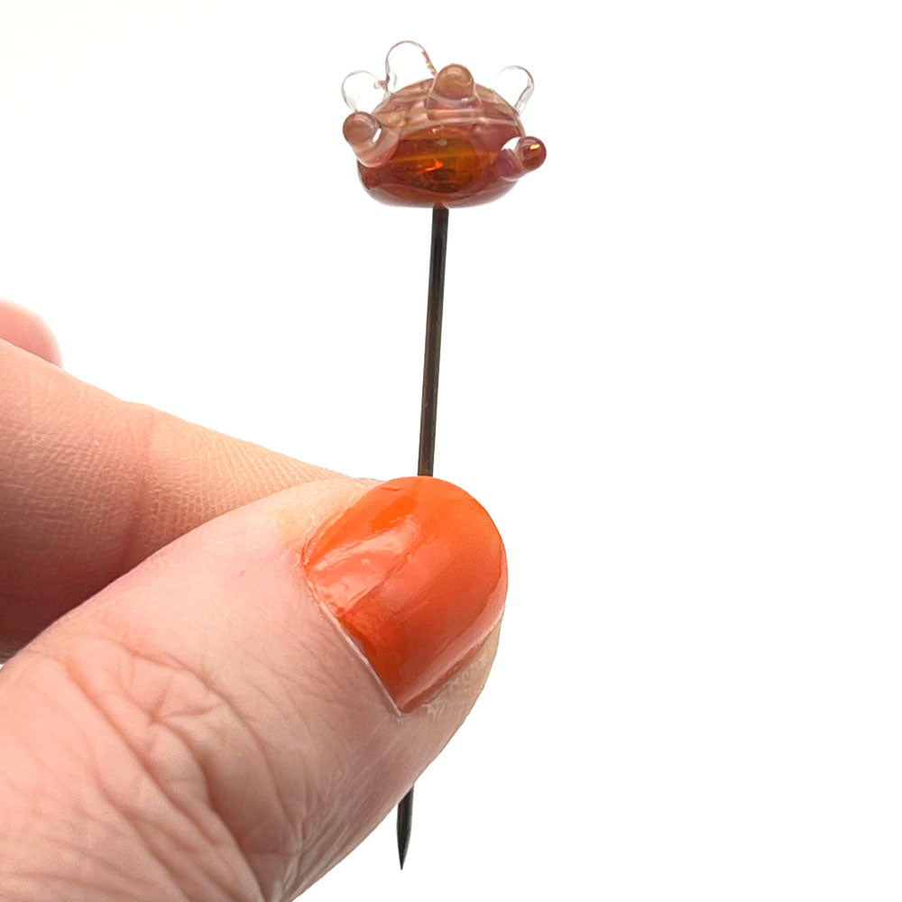 Metal pin with glass sputnik top held between thumb and forefinger. The sputnik is a  ball of swirly orange glass with raised clear dots.