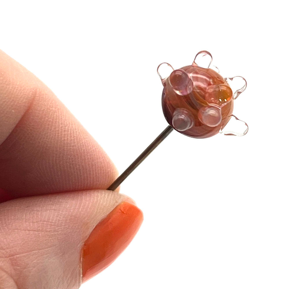 Glass headed pin. The decoration is a sputnik - a globe of swirly orange glass with raised  clear dots. Shown held between a thumb and forefinger.