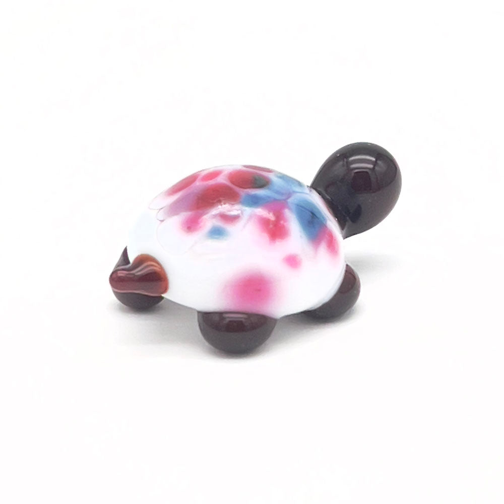 A small, glass tortoise with a white shell decorated with pink and blue speckles. The tortoise has a dark brown head and legs, displayed on a white background.