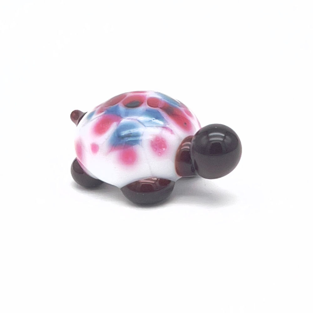 The front view of the glass tortoise, highlighting its white shell with pink and blue speckles, dark brown head, and legs, positioned on a white background.