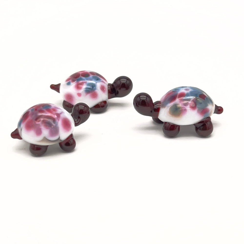 A group of three similar glass tortoises, each with a white shell and pink and blue speckles, dark brown heads, and legs, arranged on a white background.