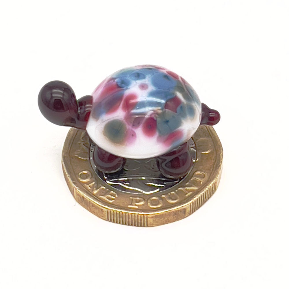 The miniature glass tortoise sitting on a one-pound coin, highlighting its small size. The tortoise has a white shell with pink and blue speckles, and dark brown head and legs.