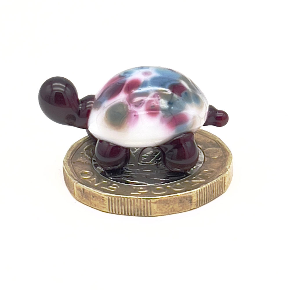 The glass tortoise on top of a one-pound coin, viewed from a different angle, emphasizing its small size. The tortoise features a white shell with pink and blue speckles and dark brown head and legs.