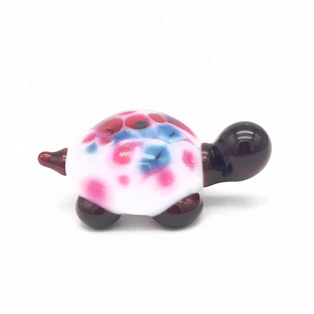 The side view of the glass tortoise, showcasing its white shell with pink and blue speckles, dark brown head, and legs, displayed on a white background.