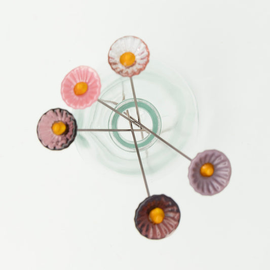 Top down view. Five purple glass flowers with metal plant stakes. Flowers are different shades of purple and have yellow centres.  The plant stakes sit in a clear milk bottle vase.