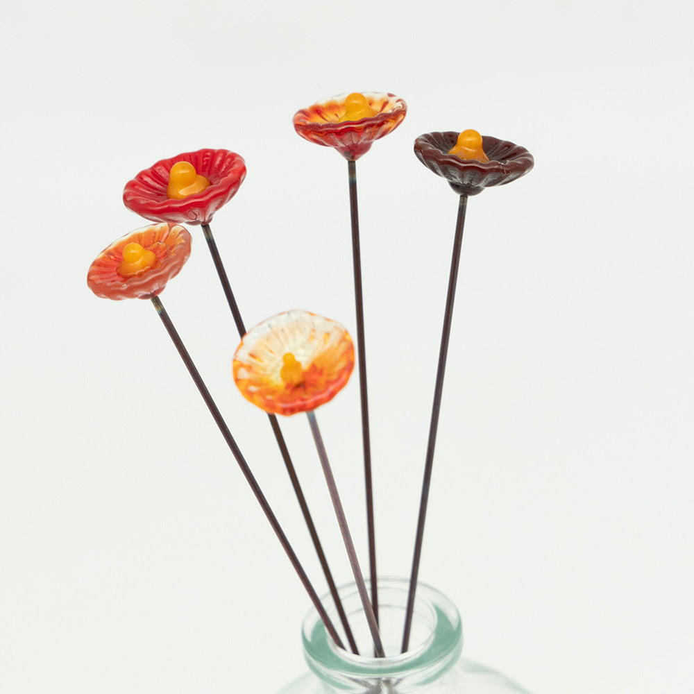 Closeup  view. Five red glass flowers with metal plant stakes. Flowers are different shades of red colours and have yellow centres.  The plant stakes sit in a clear milk bottle vase.