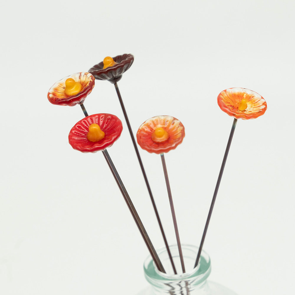 Closeup  view. Five red glass flowers with metal plant stakes. Flowers are different shades of red colours and have yellow centres.  The plant stakes sit in a clear milk bottle vase.