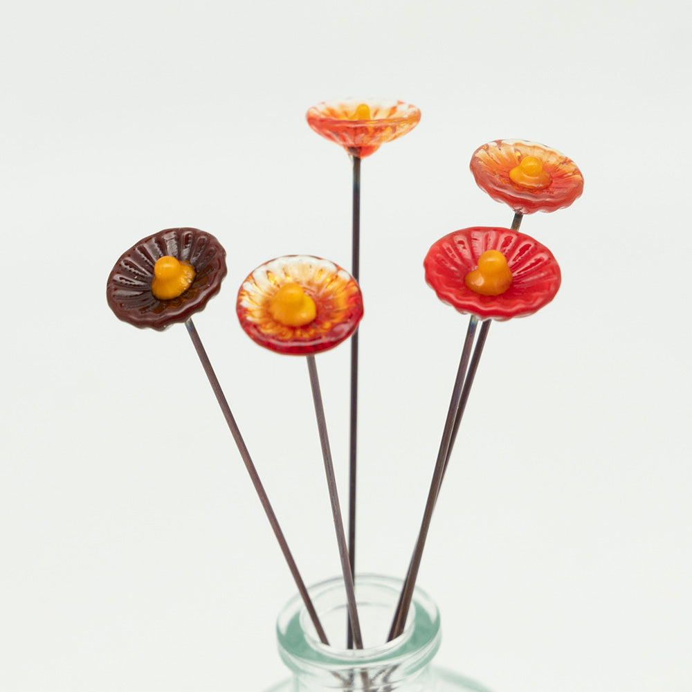 Closeup  view. Five red glass flowers with metal plant stakes. Flowers are different shades of red colours and have yellow centres.  The plant stakes sit in a clear milk bottle vase.