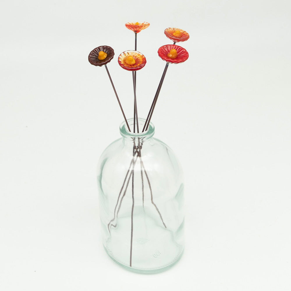 Five red glass flowers with metal plant stakes. Flowers are different shades of red colours and have yellow centres.  The plant stakes sit in a clear milk bottle vase.