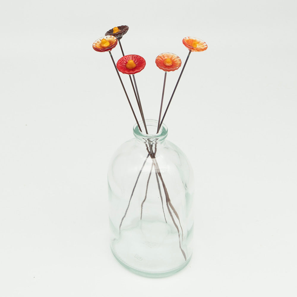 Five red glass flowers with metal plant stakes. Flowers are different shades of red colours and have yellow centres.  The plant stakes sit in a clear milk bottle vase.