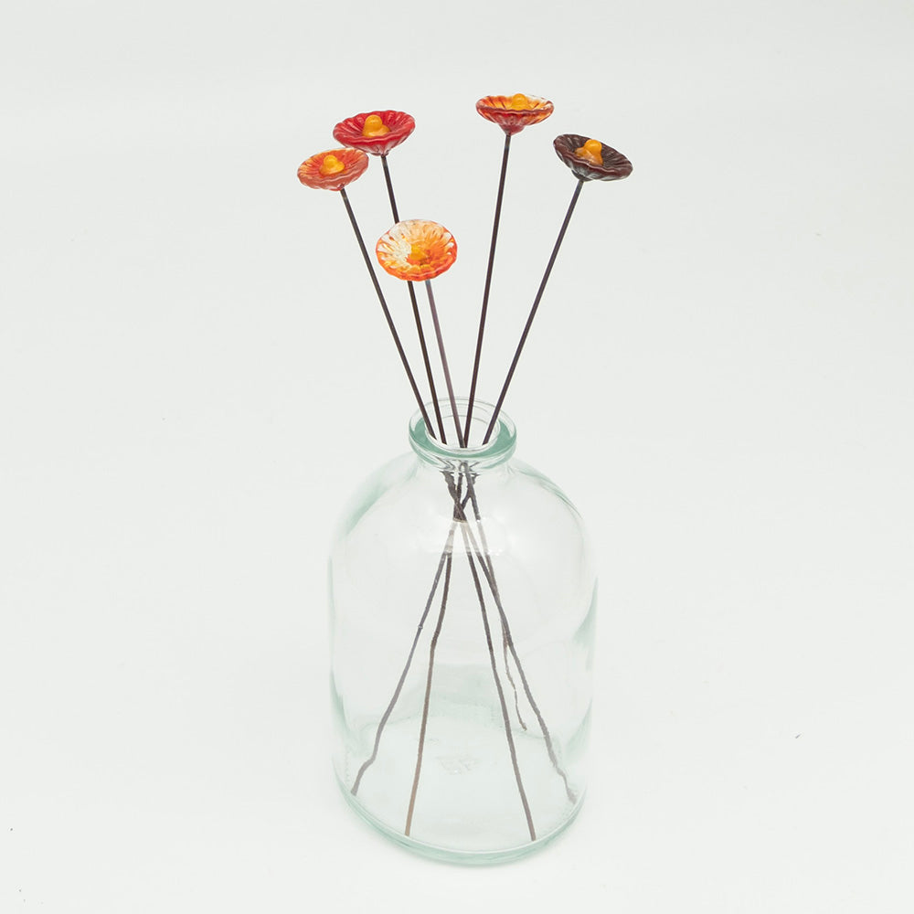 Five red glass flowers with metal plant stakes. Flowers are different shades of red colours and have yellow centres.  The plant stakes sit in a clear milk bottle vase.