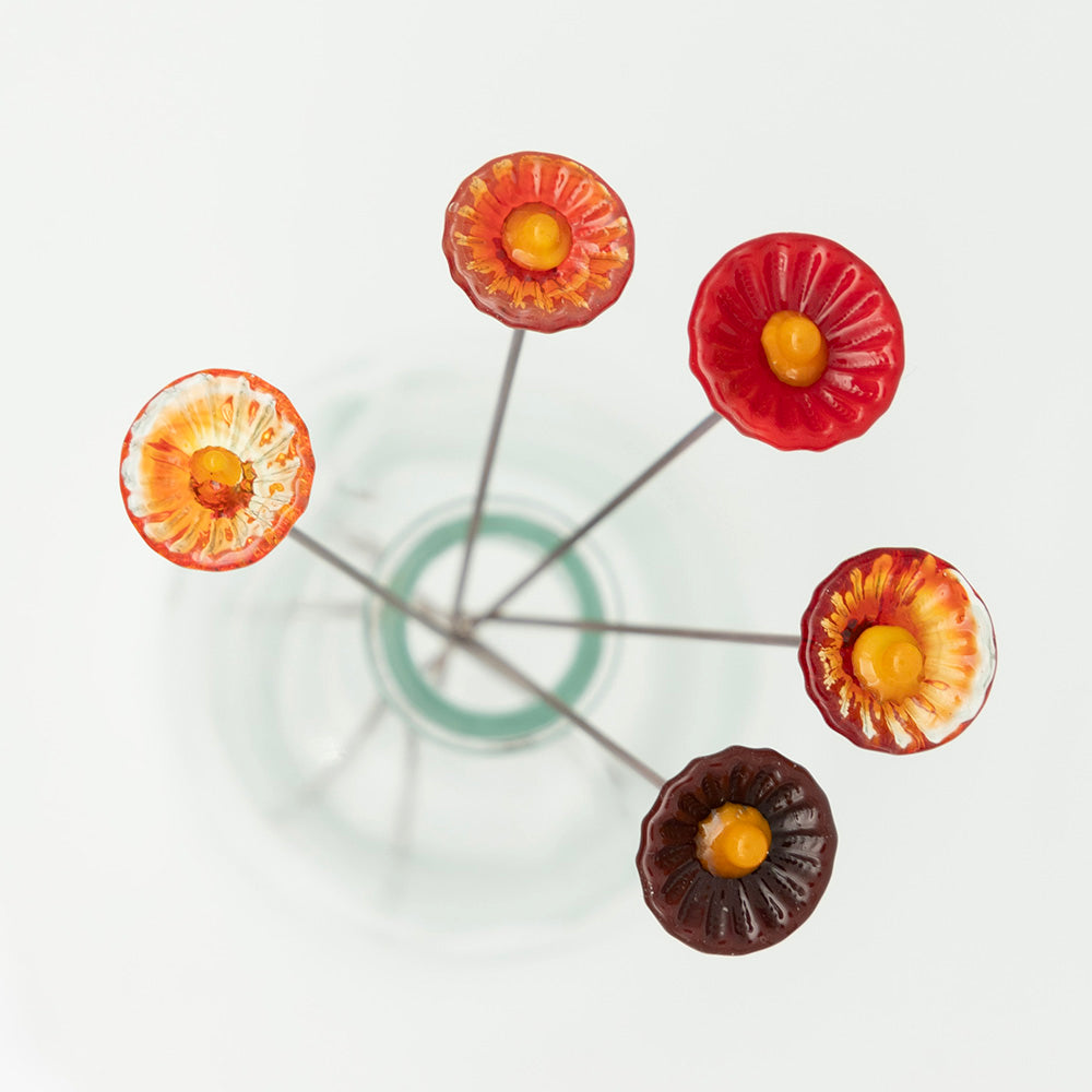 Overhead view. Five red glass flowers with metal plant stakes. Flowers are different shades of red colours and have yellow centres.  The plant stakes sit in a clear milk bottle vase.