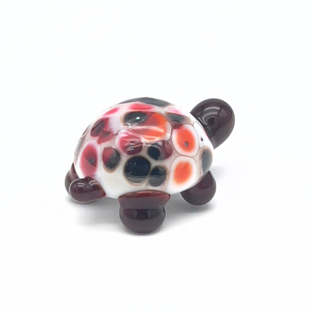 A small, glass tortoise with a white shell decorated with red, orange, and black speckles. The tortoise has a dark brown head and legs, displayed on a white background.