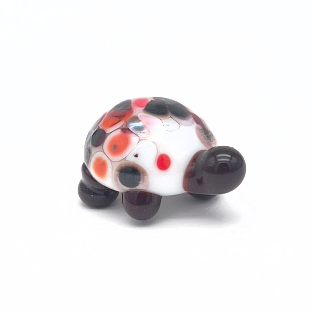 The front view of the glass tortoise, highlighting its white shell with red, orange, and black speckles, dark brown head, and legs, positioned on a white background.