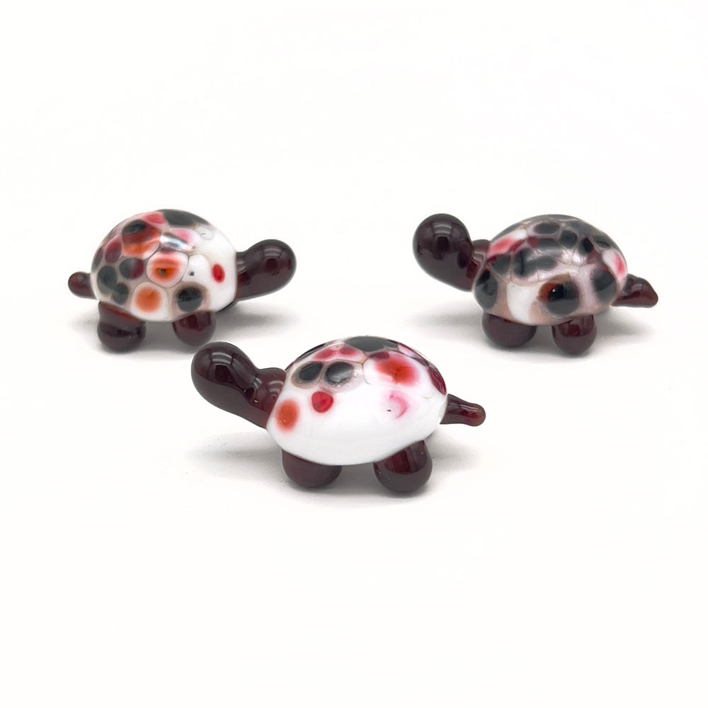 A group of three similar glass tortoises, each with a white shell and red, orange, and black speckles, dark brown heads, and legs, arranged on a white background.