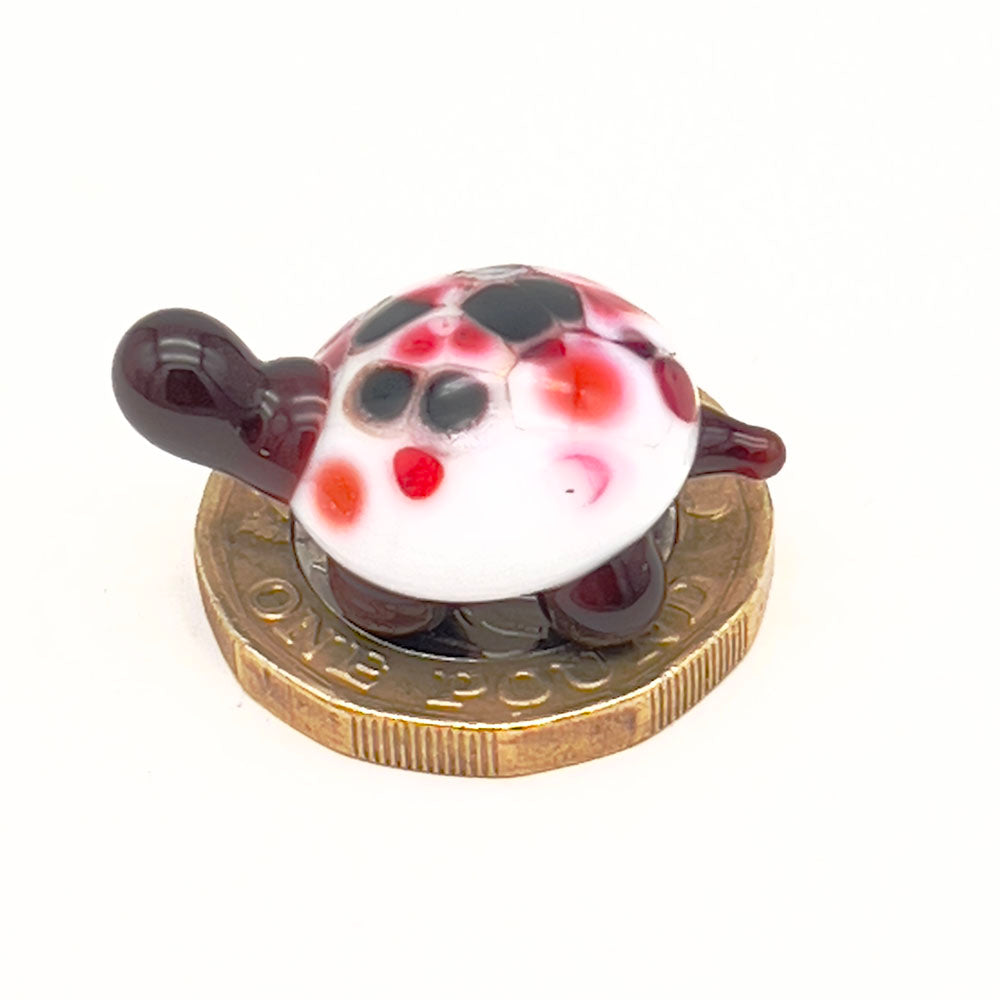 The miniature glass tortoise sitting on a one-pound coin, highlighting its small size. The tortoise has a white shell with red, orange, and black speckles, and dark brown head and legs.