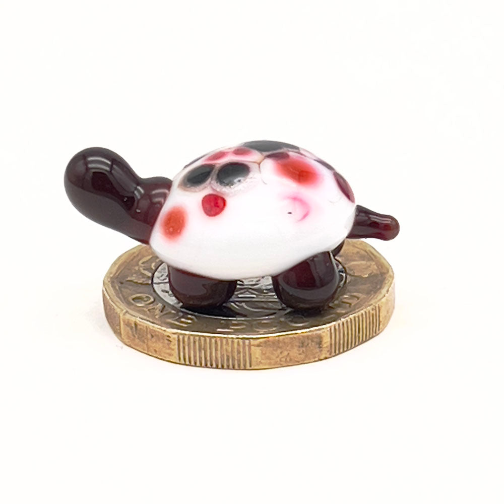 The glass tortoise on top of a one-pound coin, viewed from a different angle, emphasizing its small size. The tortoise features a white shell with red, orange, and black speckles and dark brown head and legs.