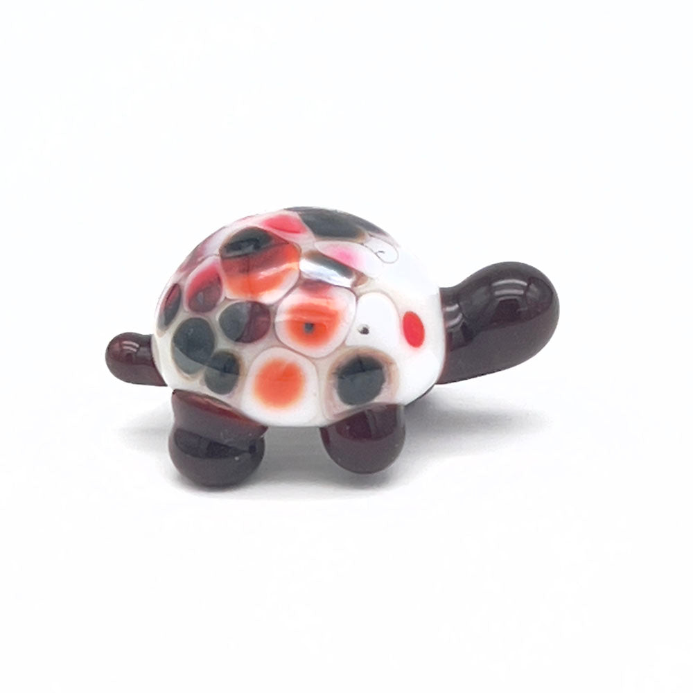 The side view of the glass tortoise, showcasing its white shell with red, orange, and black speckles, dark brown head, and legs, displayed on a white background.