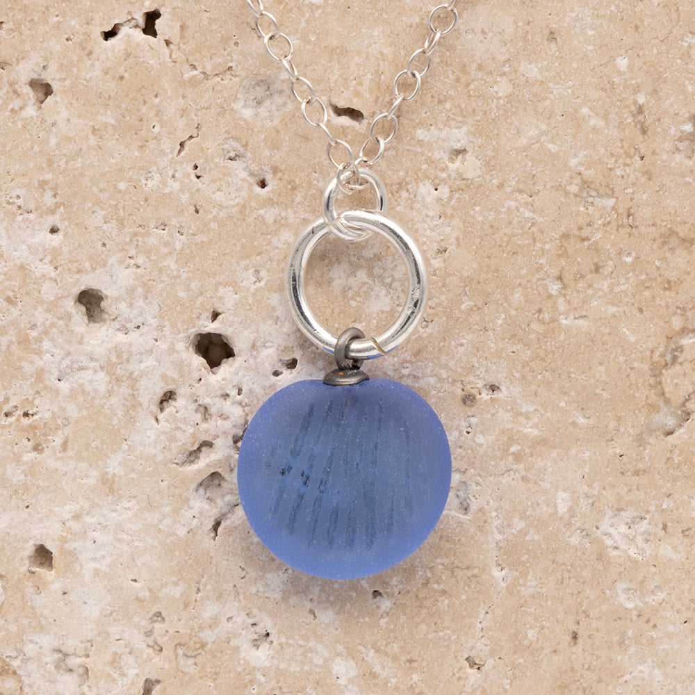 Close up of the front of a blue frosted glass charm with shell imprint on a sandstone background. The charm is frosted, and spiral is shiny. The charm hangs on a large silver jump ring. The necklace is completed with a small silver jump ring and silver chain.