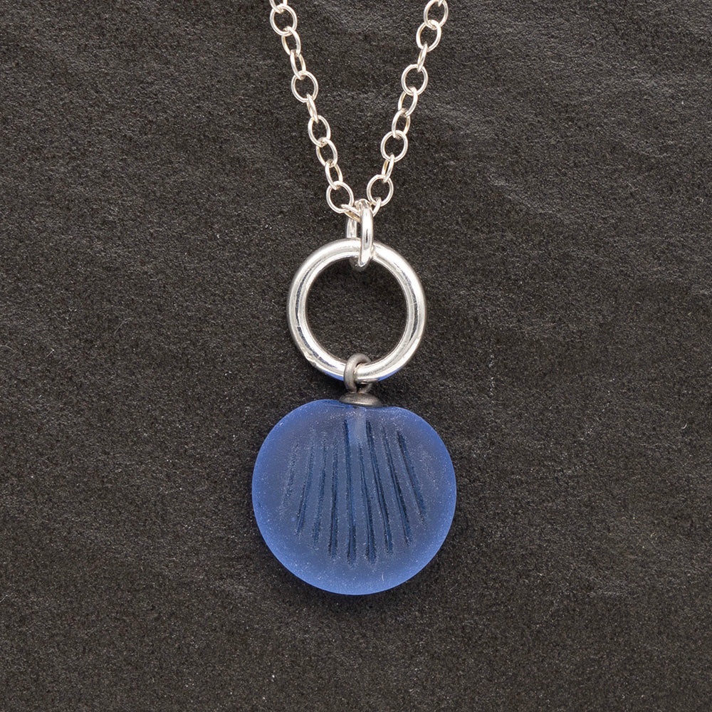 Close up of a blue frosted glass charm with shell imprint on a dark slate background. The charm is frosted, and spiral is shiny. The charm hangs on a large silver jump ring. The necklace is completed with a small silver jump ring and silver chain.