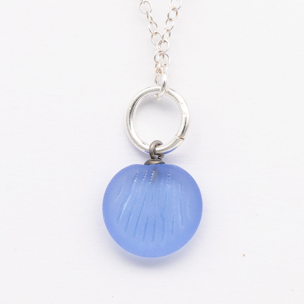 Close up of blue frosted glass charm with shell imprint on a plain white background. The charm is frosted, and spiral is shiny. The charm hangs on a large silver jump ring. The necklace is completed with a small silver jump ring and silver chain.