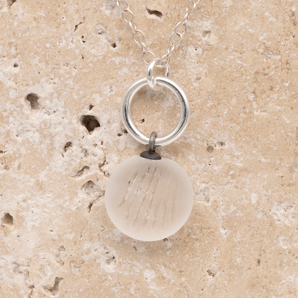 Close up of the front of a clear frosted glass charm with shell imprint on a sandstone background. The charm is frosted, and spiral is shiny. The charm hangs on a large silver jump ring. The necklace is completed with a small silver jump ring and silver chain.