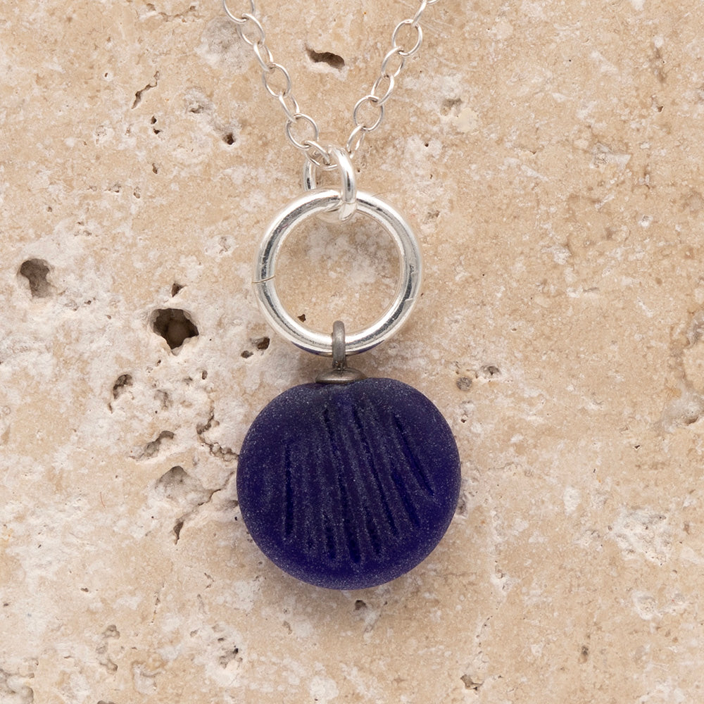 Close up of the front of a dark blue frosted glass charm with shell imprint on a sandstone background. The charm is frosted, and spiral is shiny. The charm hangs on a large silver jump ring. The necklace is completed with a small silver jump ring and silver chain.