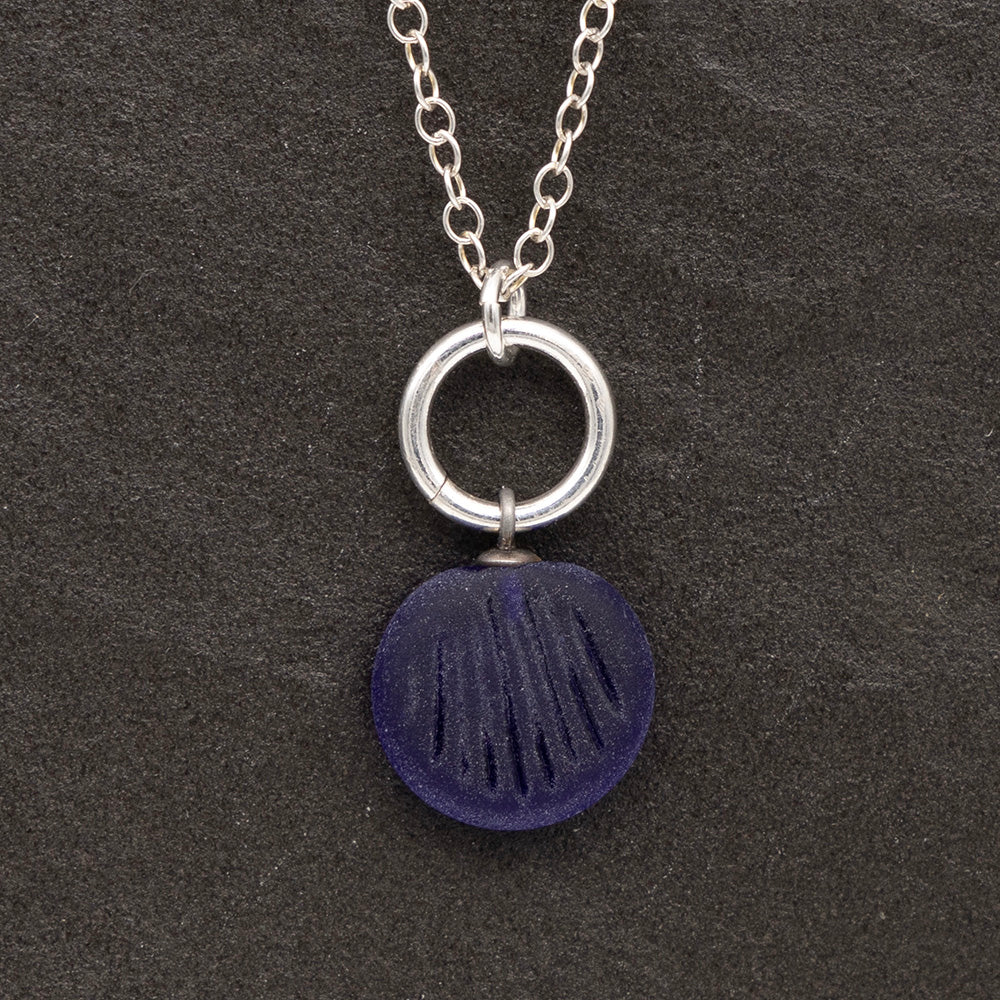 Close up of a dark blue frosted glass charm with shell imprint on a dark slate background. The charm is frosted, and spiral is shiny. The charm hangs on a large silver jump ring. The necklace is completed with a small silver jump ring and silver chain.