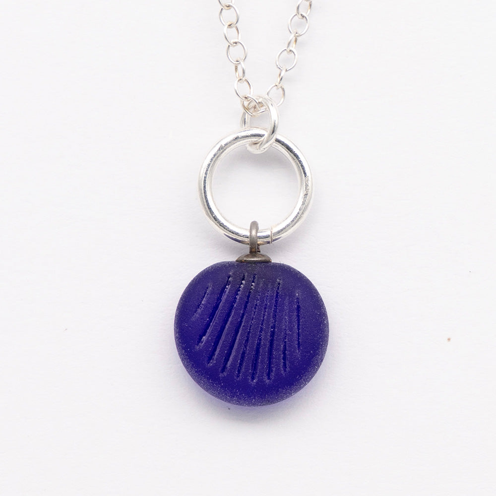 Close up of dark blue frosted glass charm with shell imprint on a plain white background. The charm is frosted, and spiral is shiny. The charm hangs on a large silver jump ring. The necklace is completed with a small silver jump ring and silver chain.