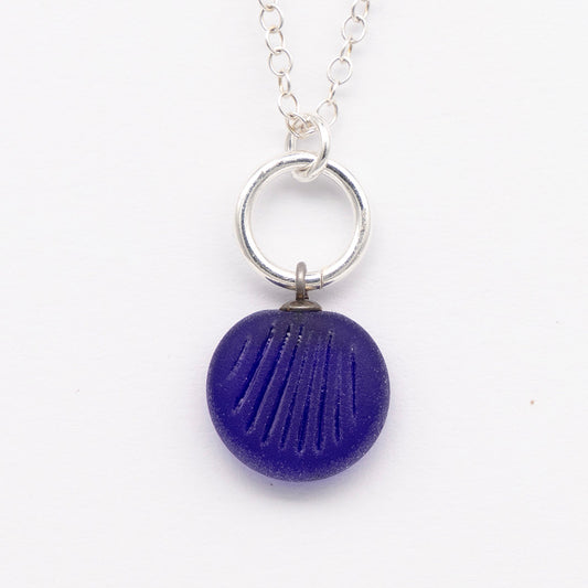 Close up of dark blue frosted glass charm with shell imprint on a plain white background. The charm is frosted, and spiral is shiny. The charm hangs on a large silver jump ring. The necklace is completed with a small silver jump ring and silver chain.