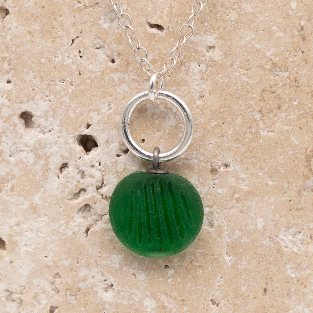 Close up of the front of a green frosted glass charm with shell imprint on a sandstone background. The charm is frosted, and spiral is shiny. The charm hangs on a large silver jump ring. The necklace is completed with a small silver jump ring and silver chain.