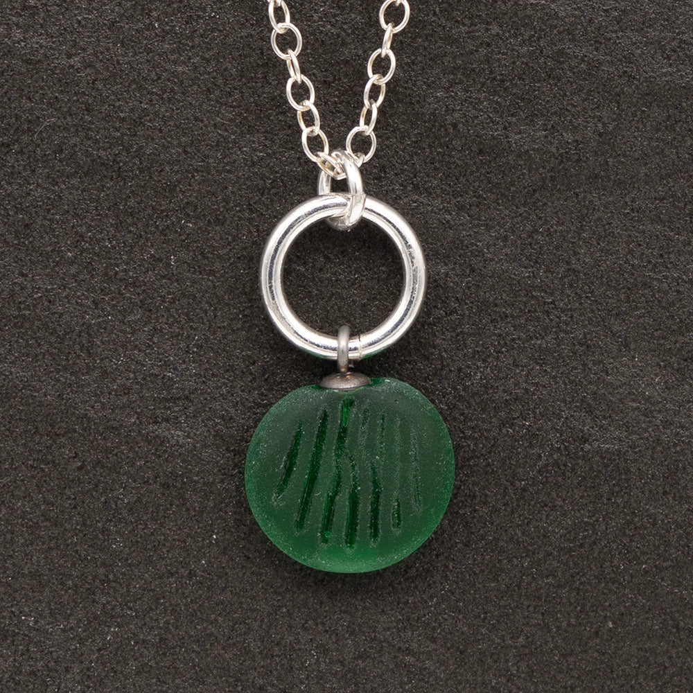 Close up of a green frosted glass charm with shell imprint on a dark slate background. The charm is frosted, and spiral is shiny. The charm hangs on a large silver jump ring. The necklace is completed with a small silver jump ring and silver chain.