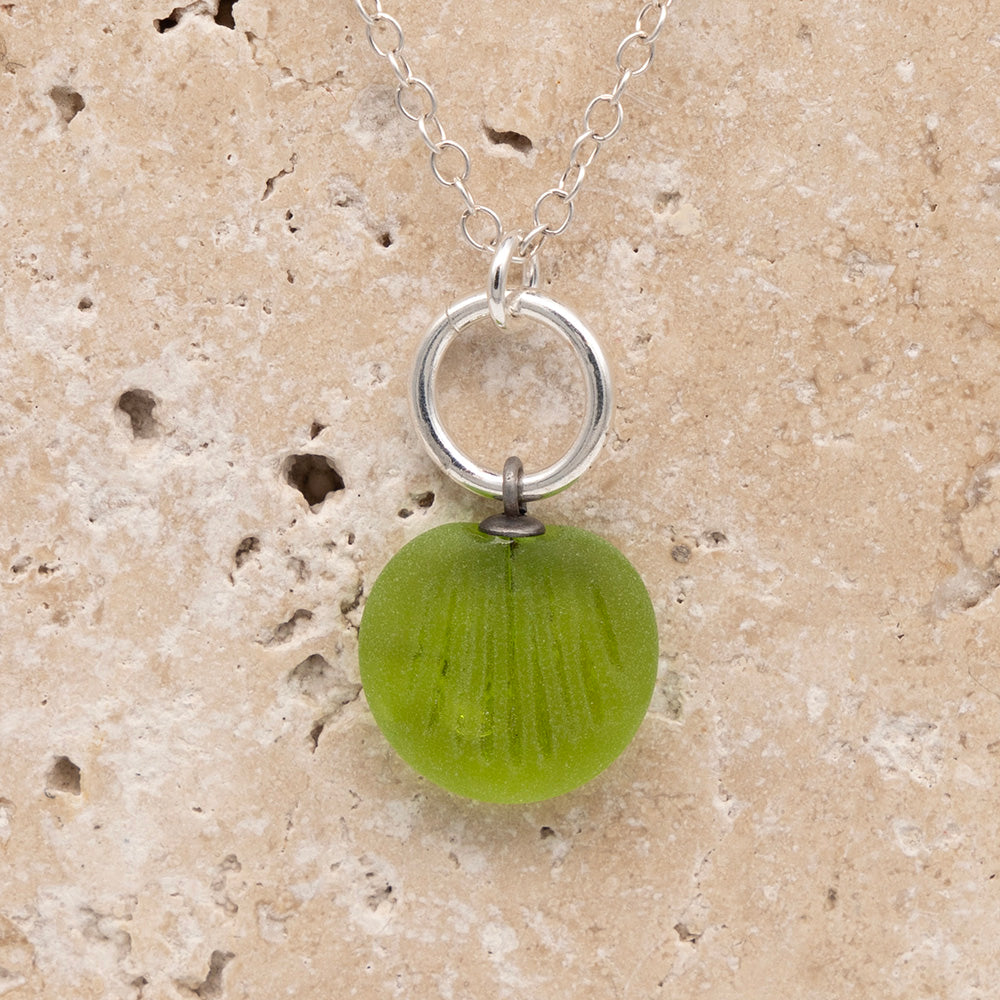 Close up of the front of a lime green frosted glass charm with shell imprint on a sandstone background. The charm is frosted, and spiral is shiny. The charm hangs on a large silver jump ring. The necklace is completed with a small silver jump ring and silver chain.