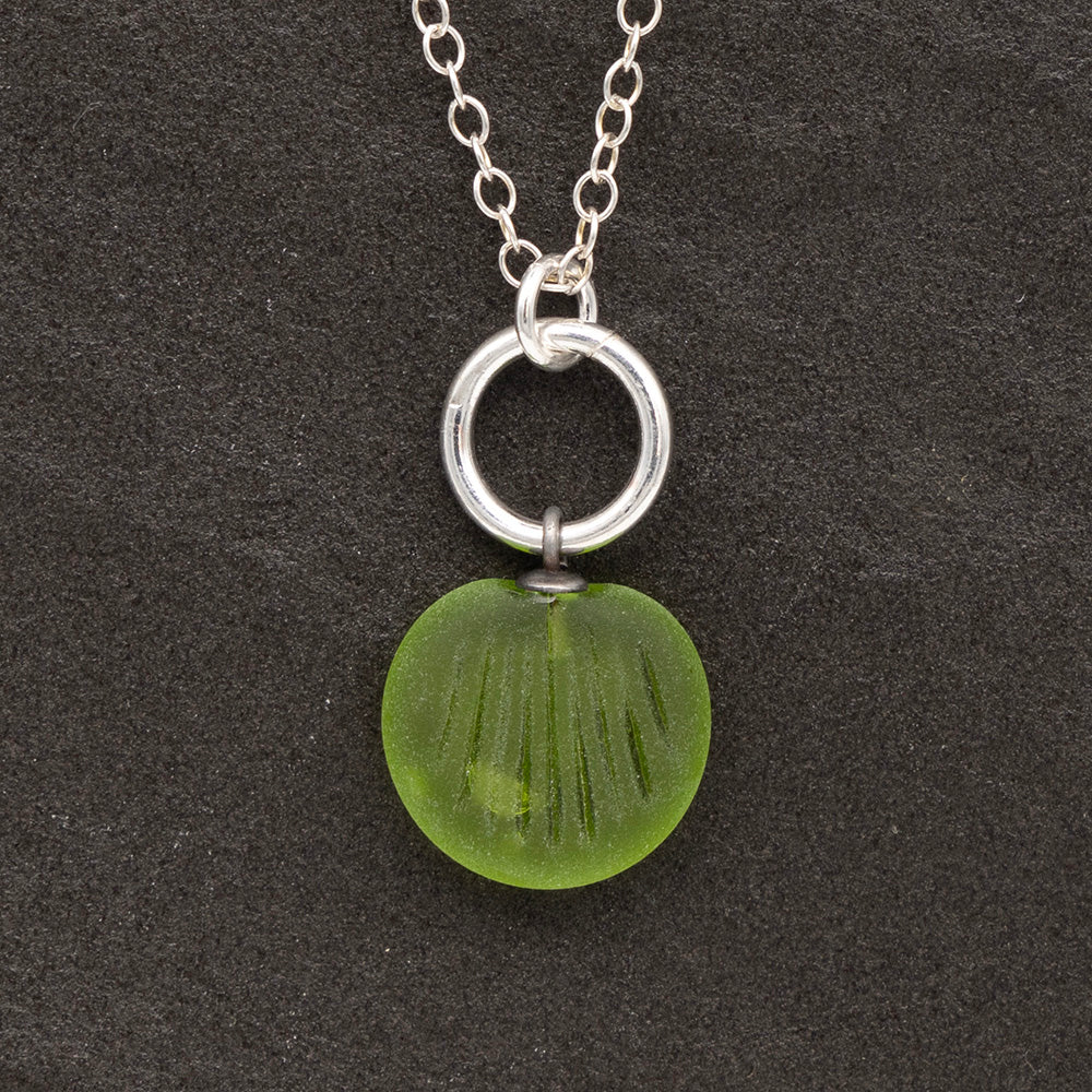 Close up of a lime green frosted glass charm with shell imprint on a dark slate background. The charm is frosted, and spiral is shiny. The charm hangs on a large silver jump ring. The necklace is completed with a small silver jump ring and silver chain.