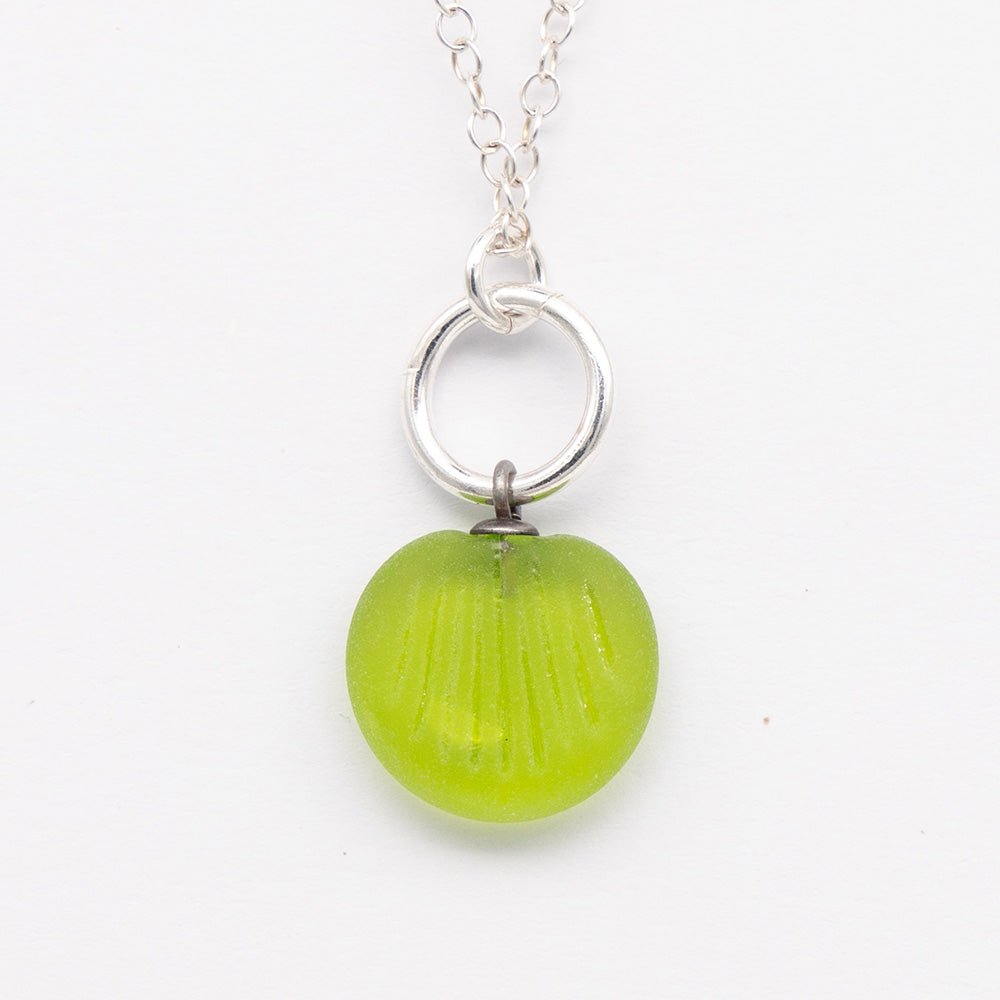 Close up of lime green frosted glass charm with shell imprint on a plain white background. The charm is frosted, and spiral is shiny. The charm hangs on a large silver jump ring. The necklace is completed with a small silver jump ring and silver chain.