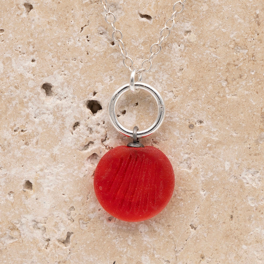 Close up of the front of an orange frosted glass charm with shell  imprint on a sandstone background. The charm is frosted, and spiral is shiny. The charm hangs on a large silver jump ring. The necklace is completed with a small silver jump ring and silver chain.