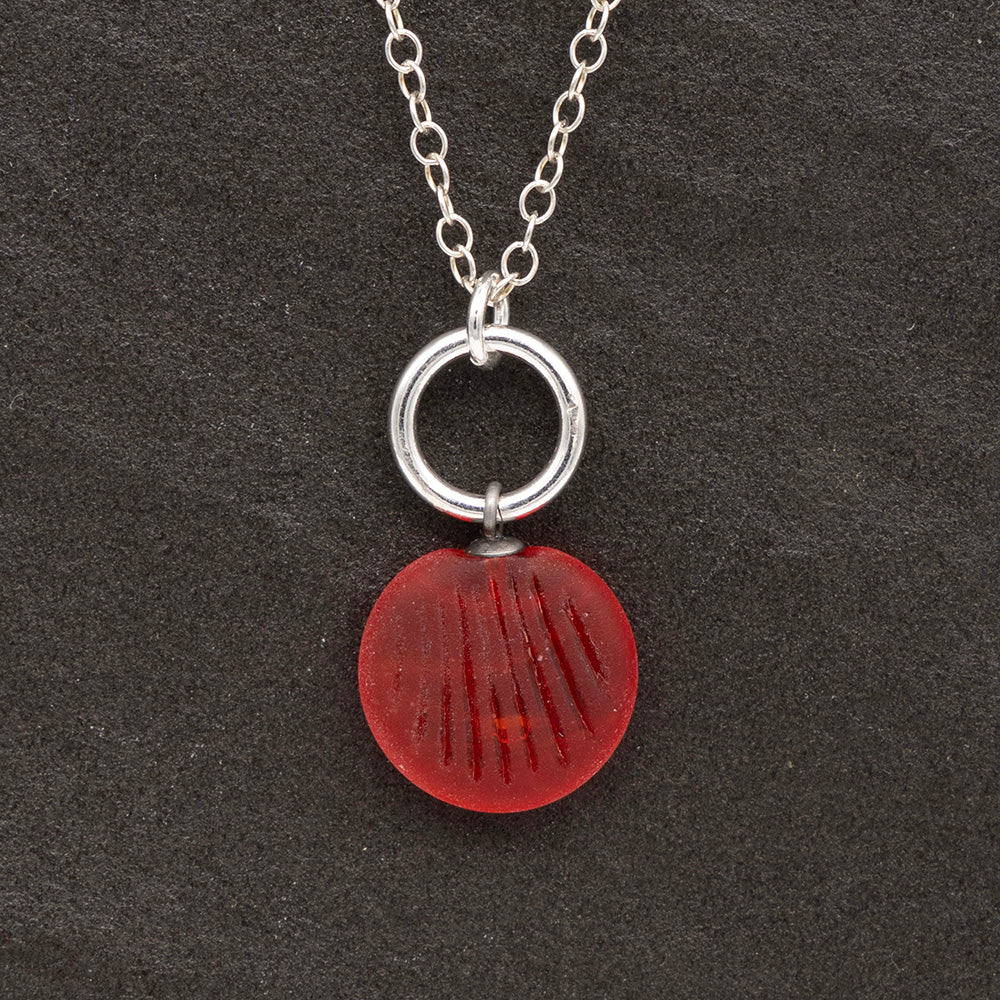 Close up of an orange frosted glass charm with shell imprint on a dark slate background. The charm is frosted, and spiral is shiny. The charm hangs on a large silver jump ring. The necklace is completed with a small silver jump ring and silver chain.