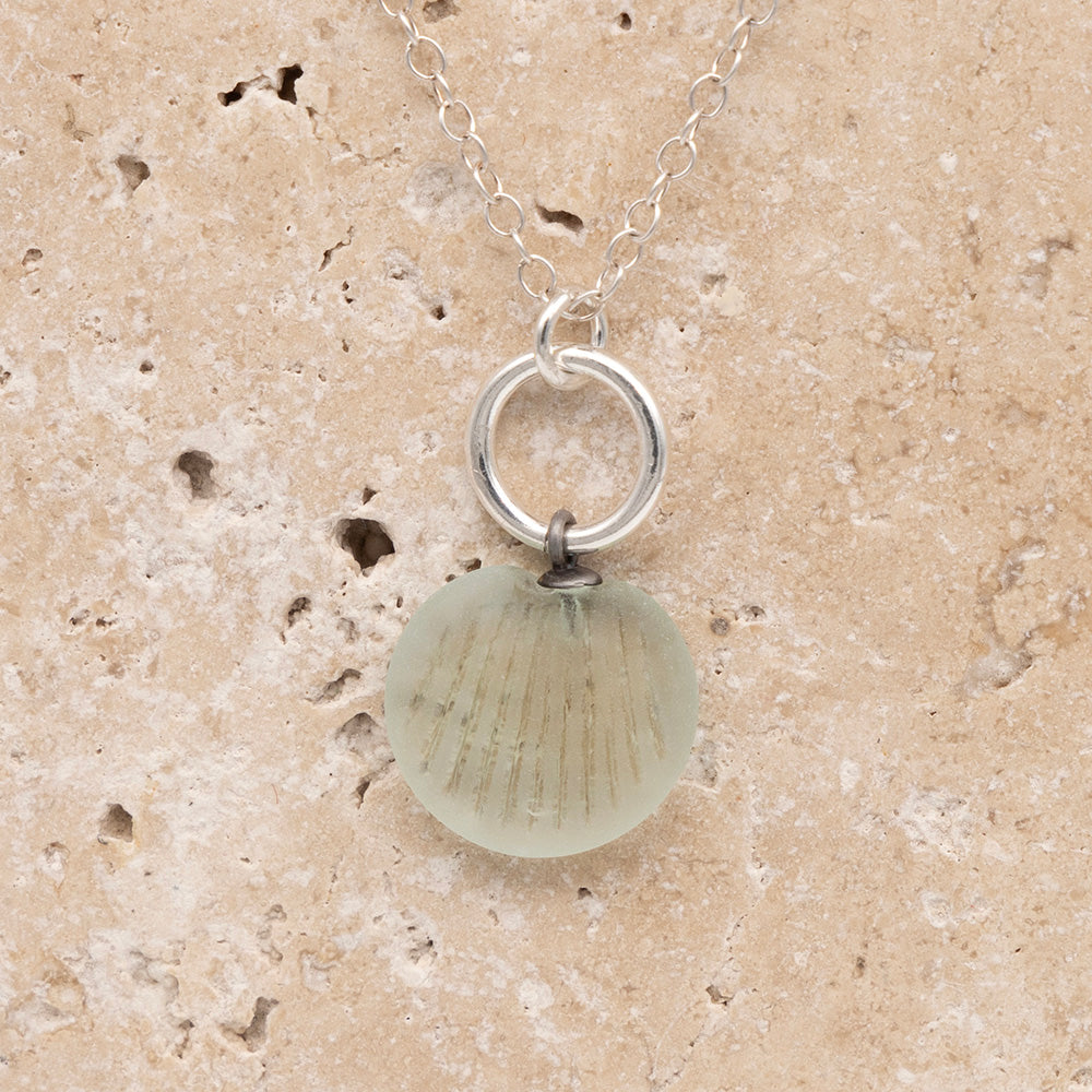 Close up of the front of a pale blue frosted glass charm with shell imprint on a sandstone background. The charm is frosted, and spiral is shiny. The charm hangs on a large silver jump ring. The necklace is completed with a small silver jump ring and silver chain.