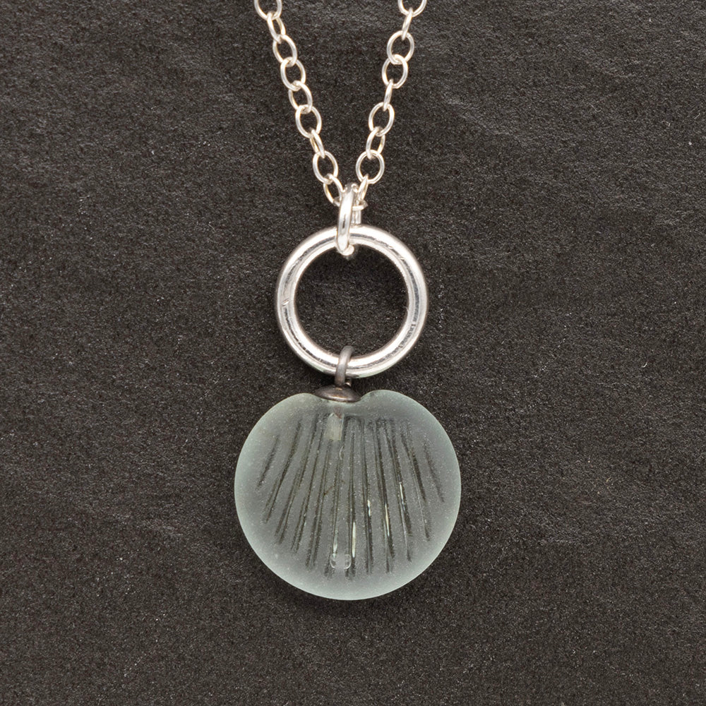 Close up of a pale blue frosted glass charm with shell imprint on a dark slate background. The charm is frosted, and spiral is shiny. The charm hangs on a large silver jump ring. The necklace is completed with a small silver jump ring and silver chain.