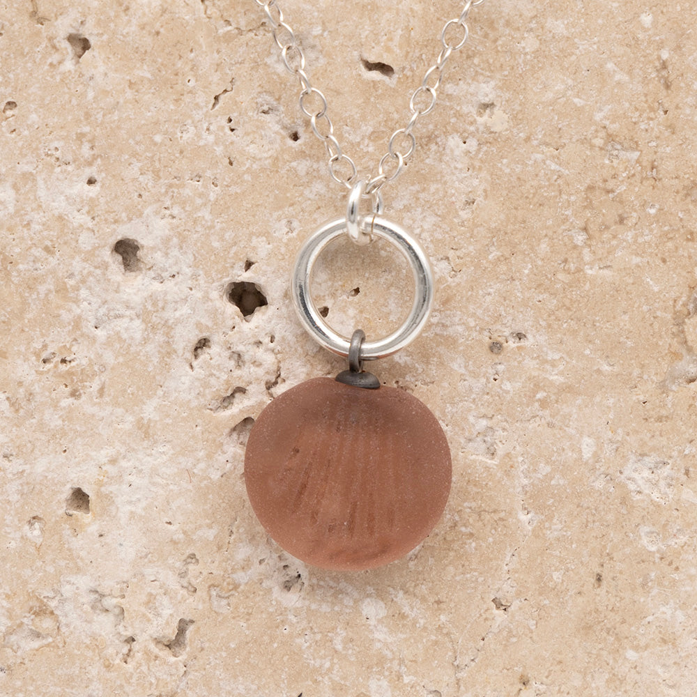 Close up of the front of a pink frosted glass charm with shell imprint on a sandstone background. The charm is frosted, and spiral is shiny. The charm hangs on a large silver jump ring. The necklace is completed with a small silver jump ring and silver chain.