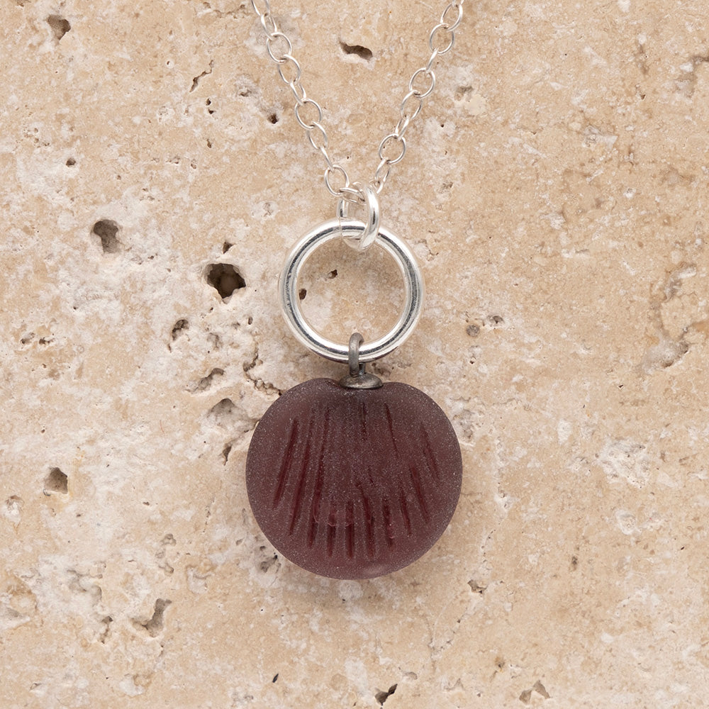 Close up of the front of a purple frosted glass charm with shell imprint on a sandstone background. The charm is frosted, and spiral is shiny. The charm hangs on a large silver jump ring. The necklace is completed with a small silver jump ring and silver chain.