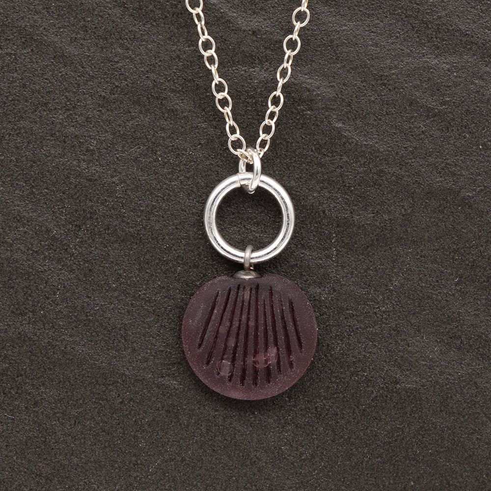 Close up of a purple frosted glass charm with shell imprint on a dark slate background. The charm is frosted, and spiral is shiny. The charm hangs on a large silver jump ring. The necklace is completed with a small silver jump ring and silver chain.