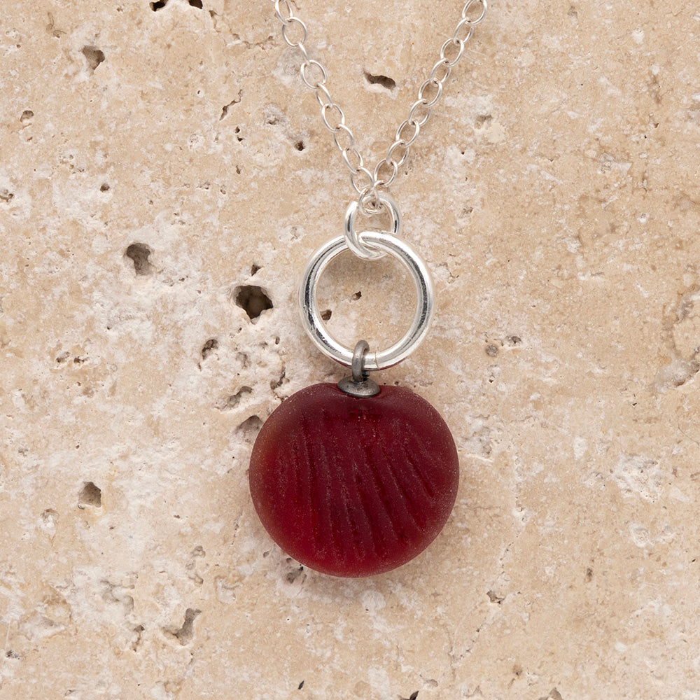 Close up of the front of a red frosted glass charm with shell imprint on a sandstone background. The charm is frosted, and spiral is shiny. The charm hangs on a large silver jump ring. The necklace is completed with a small silver jump ring and silver chain.