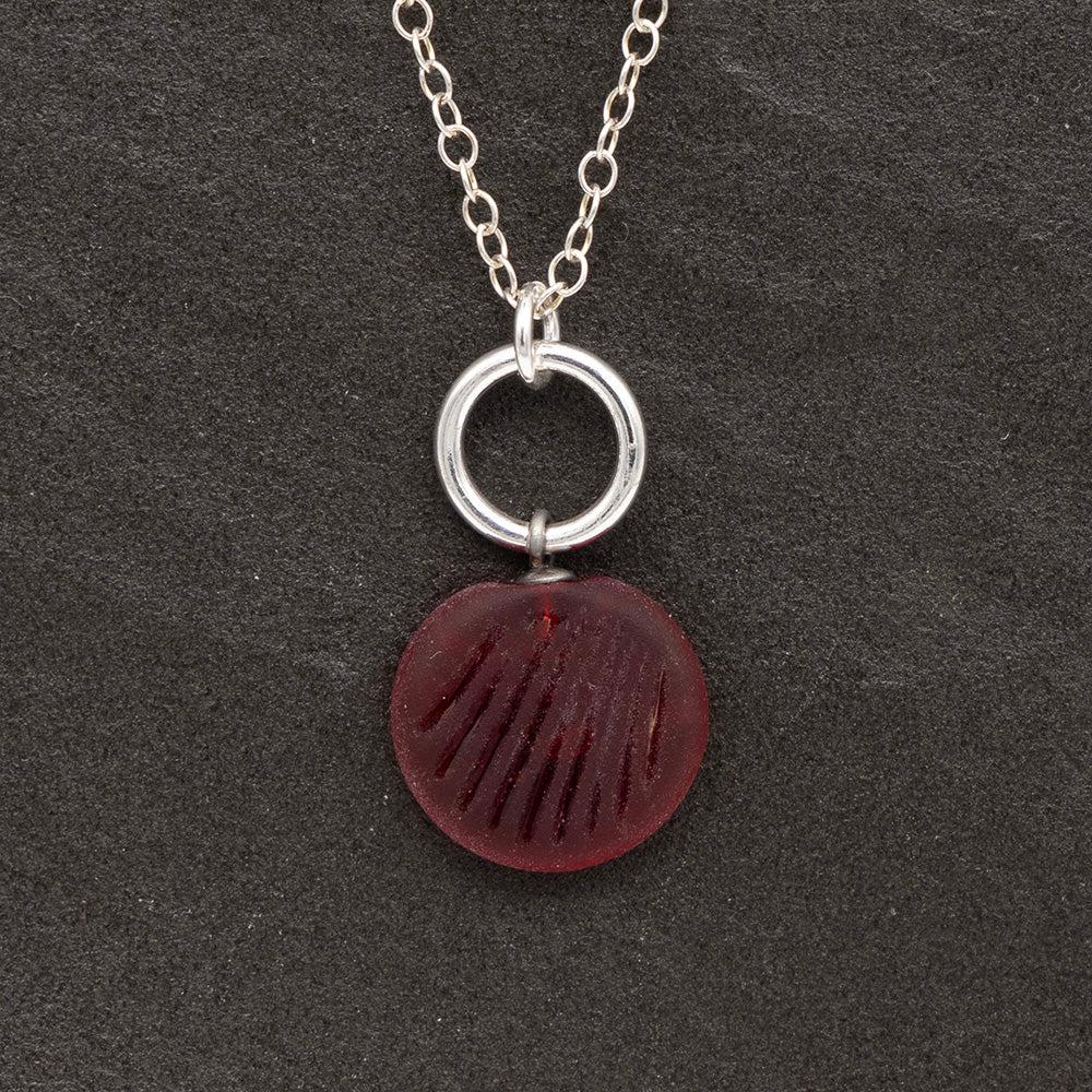 Close up of a red frosted glass charm with shell imprint on a dark slate background. The charm is frosted, and spiral is shiny. The charm hangs on a large silver jump ring. The necklace is completed with a small silver jump ring and silver chain.